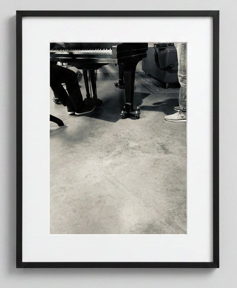 Black and white photo of a piano with people's feet near it on a concrete floor. made for music duo.
